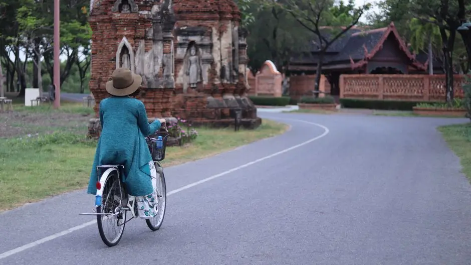 A Woman In A Hat And Cardigan Cycling Past An Ancient Temple On A Quie