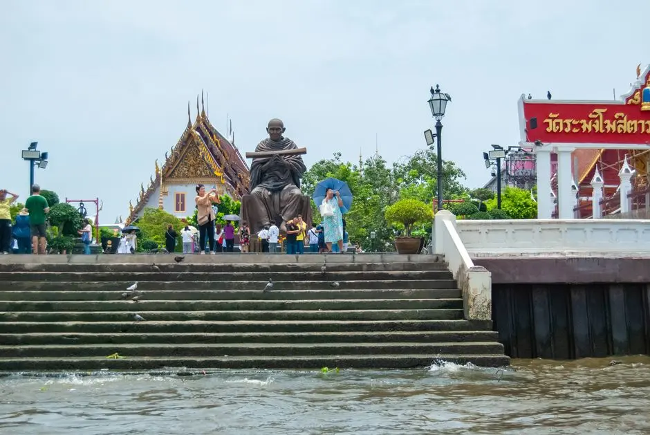 Tourists At A Riverside Statue Near A Temple In Thailand With Vibrant